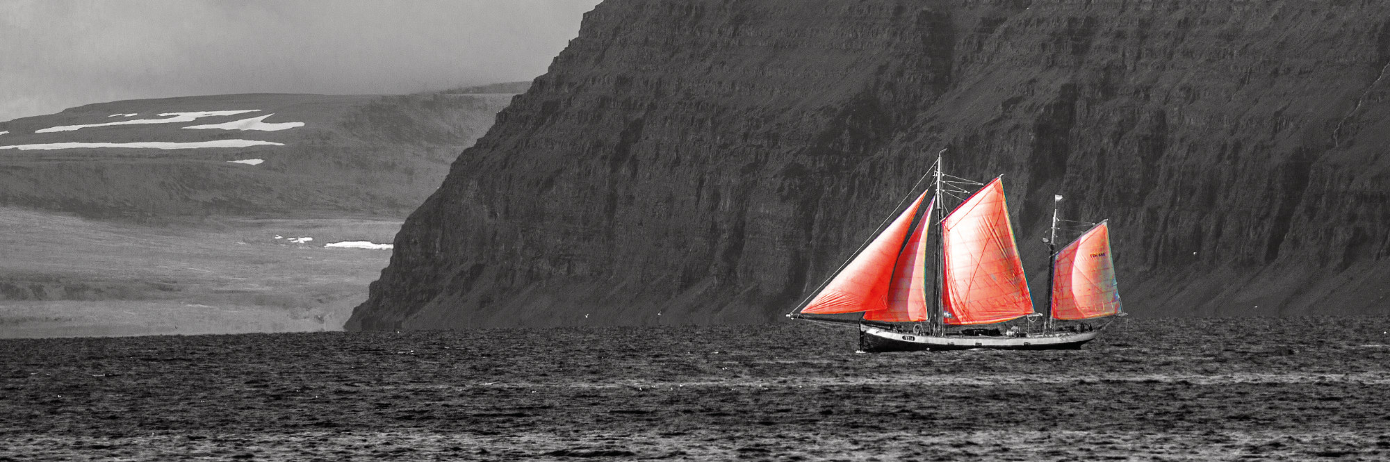 Segelschiff mit roten Segeln vor einer steilen Felsküste, Schwarzweißfoto mit roten Farbakzenten